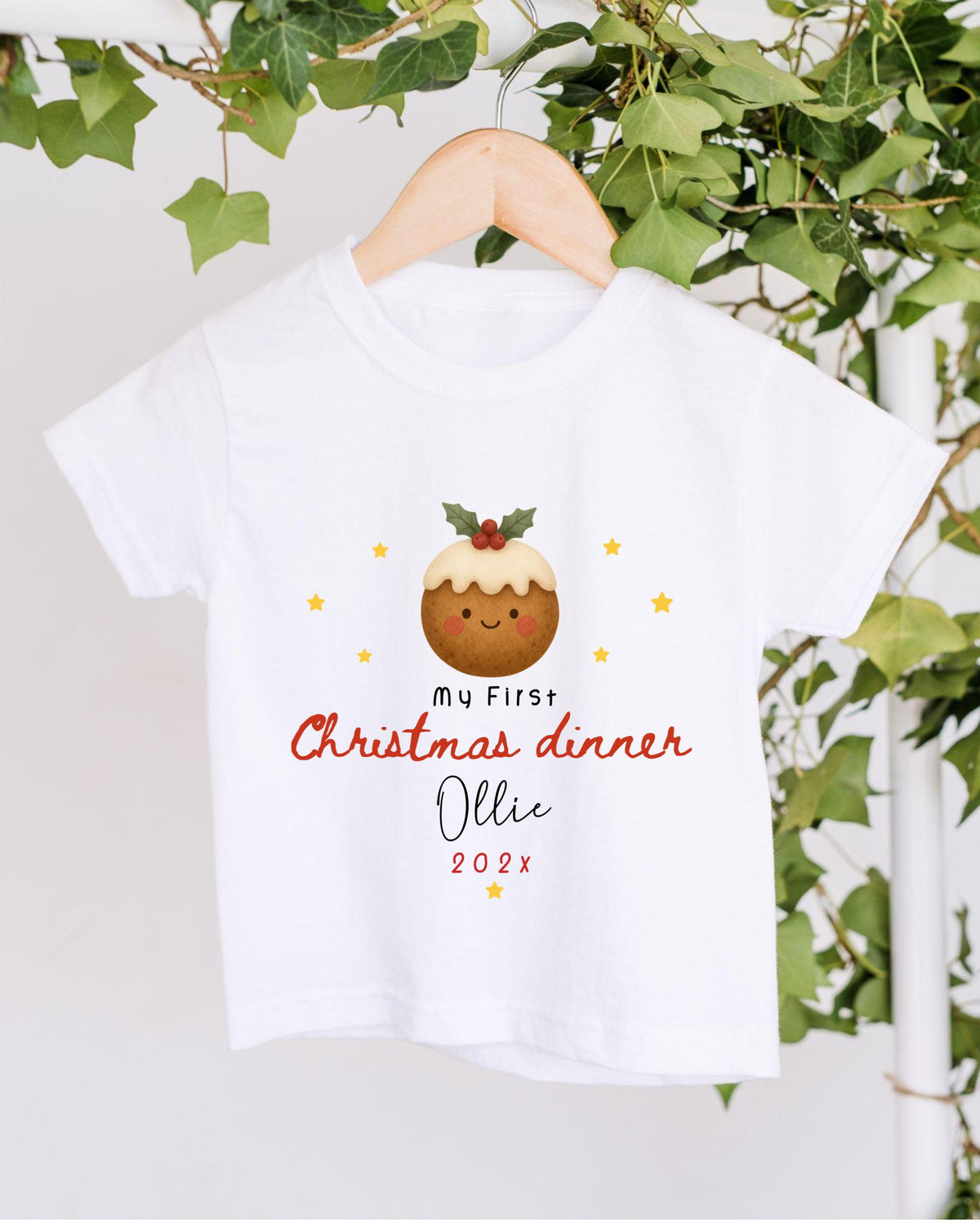 A white t-shirt with a Christmas-themed design hanging on a wooden hanger, surrounded by greenery.