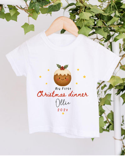 A white t-shirt with a Christmas-themed design hanging on a wooden hanger, surrounded by greenery.