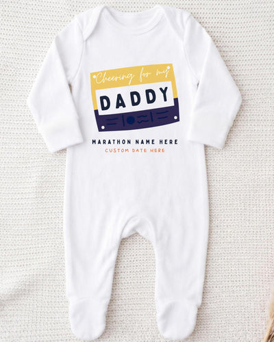 A white onesie with a graphic design on it, featuring a yellow and blue rectangle with the text "Cheering for my DADDY" and "MARCHON NAME HERE" below it.