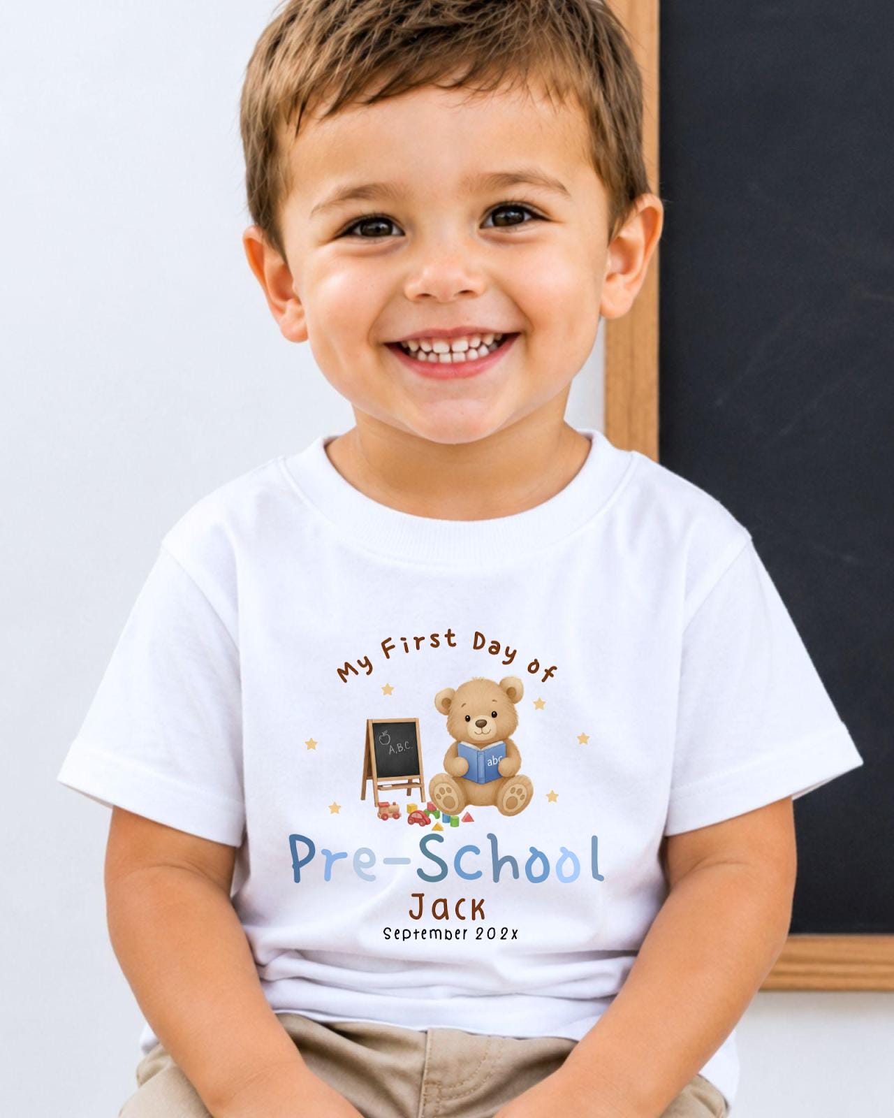A young boy wearing a white t-shirt with a teddy bear and a chalkboard, smiling at the camera.