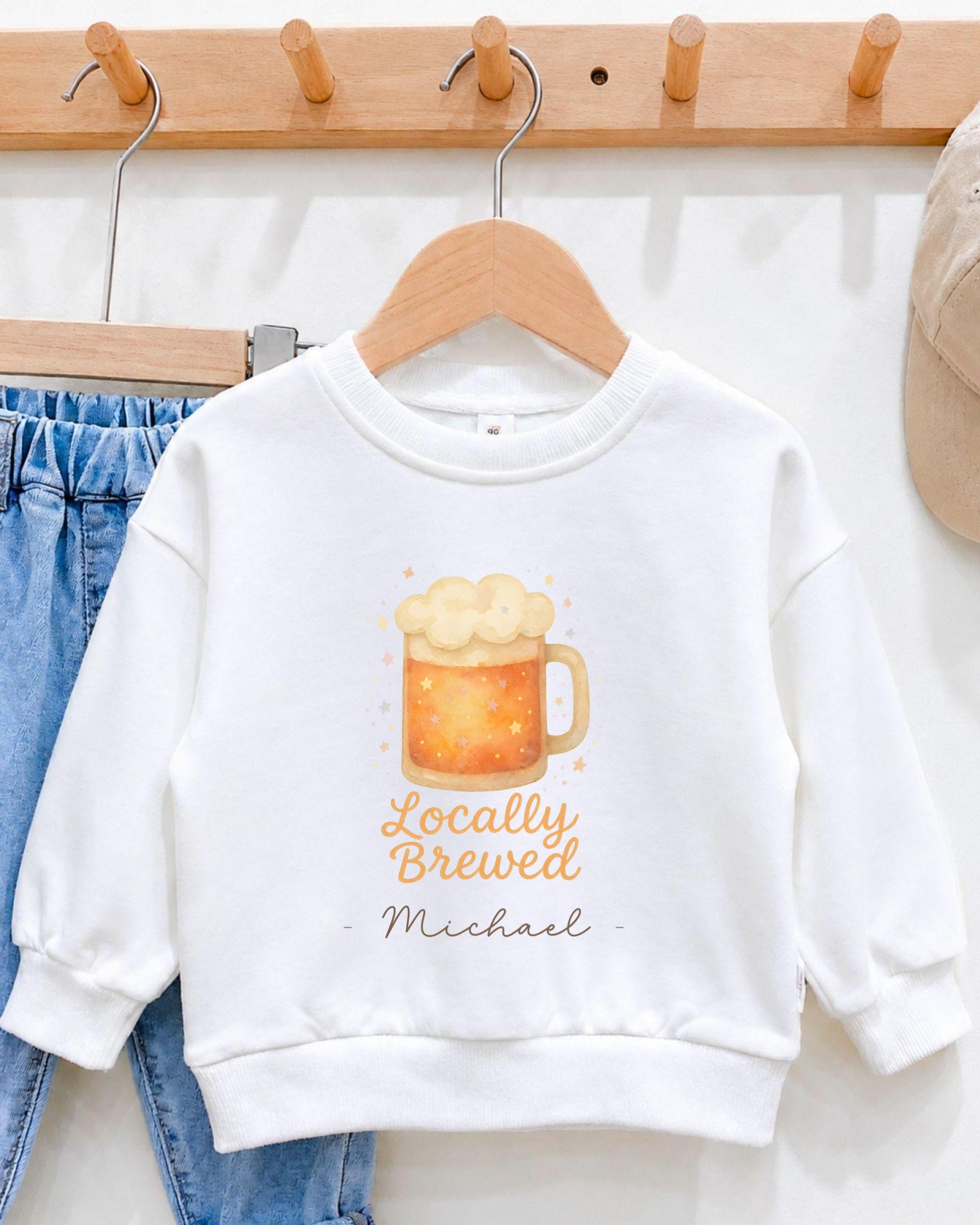 A white sweatshirt with a graphic of a mug of beer and the text "Locally Brewed Michael" printed on it, hanging on a wooden hanger.