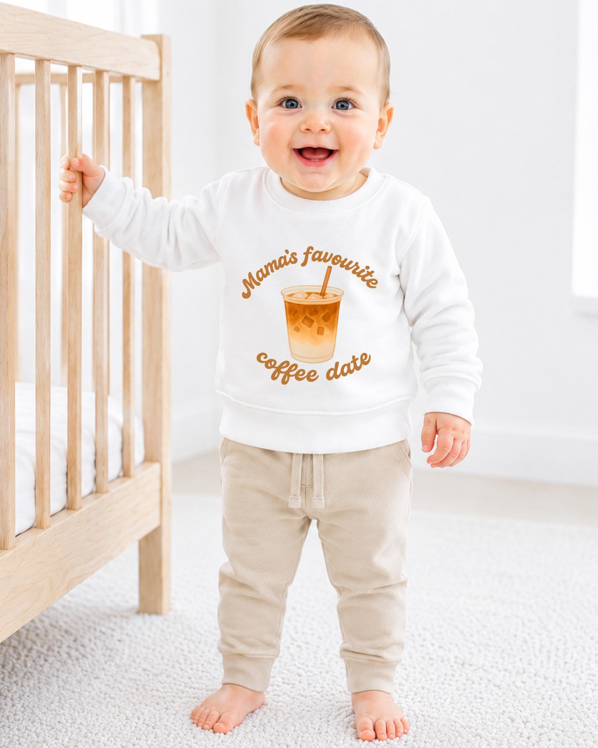 A smiling baby boy wearing a white sweatshirt with a coffee cup graphic and the text "Mama's favourite coffee date" standing in a nursery.