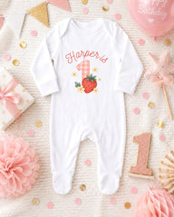 A white onesie with a strawberry and flowers printed on it, surrounded by pink and gold decorations, including a pink star, confetti, and a pink balloon with the word "Happy Birthday" written on it.
