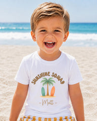 A young boy with a big smile on his face, wearing a white t-shirt with a graphic design of a palm tree and surfboards, standing on a sandy beach with the ocean in the background.