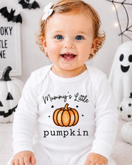 A smiling baby girl wearing a white onesie with a pumpkin and the text "Mummy's Little pumpkin" is sitting in front of Halloween decorations.