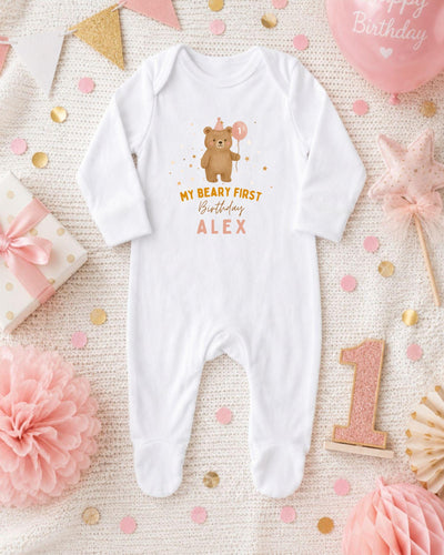 A white bodysuit with a teddy bear and the text "My first birthday Alex" printed on it, surrounded by pink and gold confetti, a pink balloon, and other baby-related items.