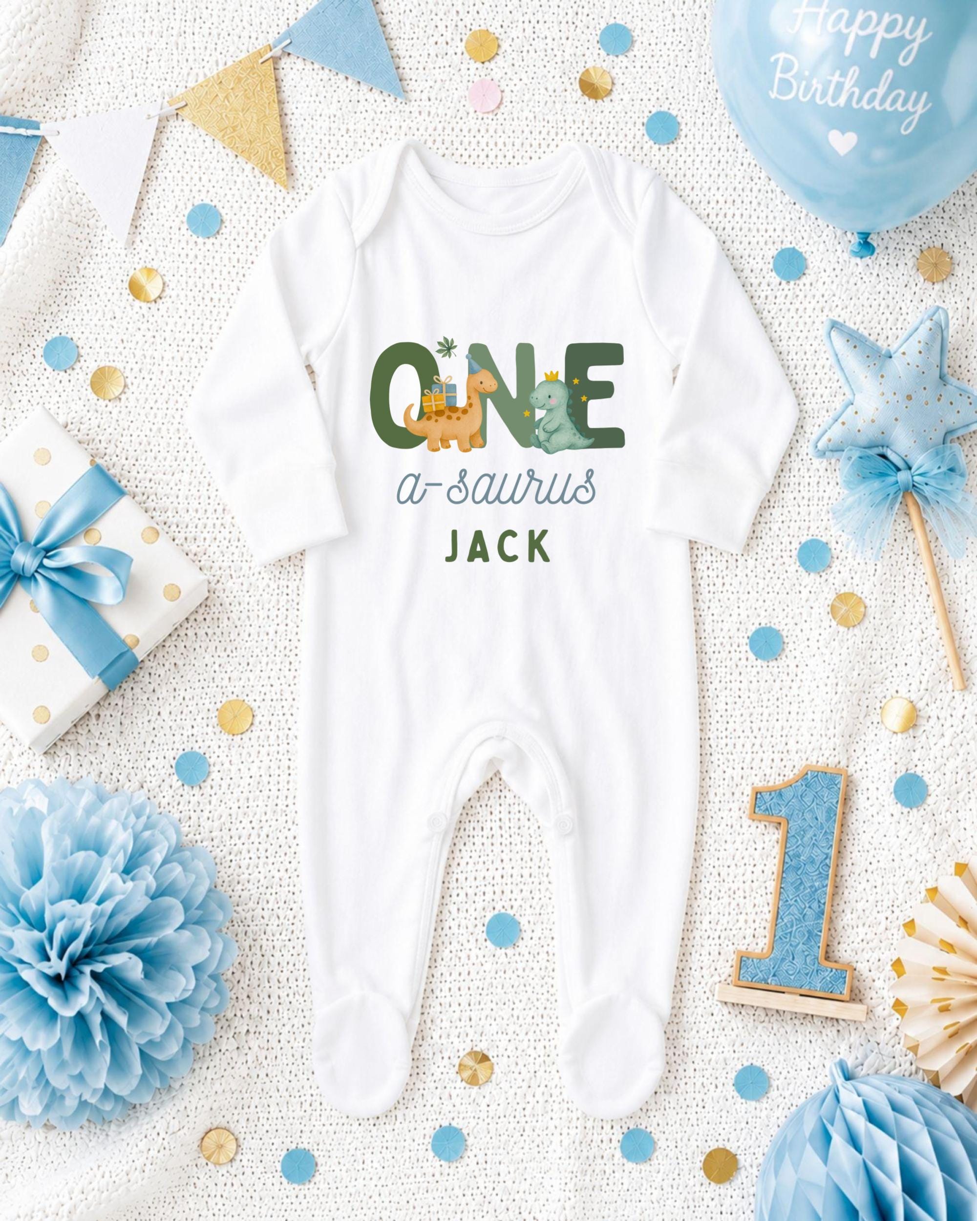 A white onesie with the text "ONE a sauropod JACK" printed on it, surrounded by various baby-themed items such as a blue balloon, a blue and white polka dot gift box, a blue and white star, a blue and white pom pom, and a blue and white confetti.