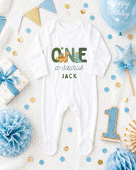 A white onesie with the text "ONE a sauropod JACK" printed on it, surrounded by various baby-themed items such as a blue balloon, a blue and white polka dot gift box, a blue and white star, a blue and white pom pom, and a blue and white confetti.