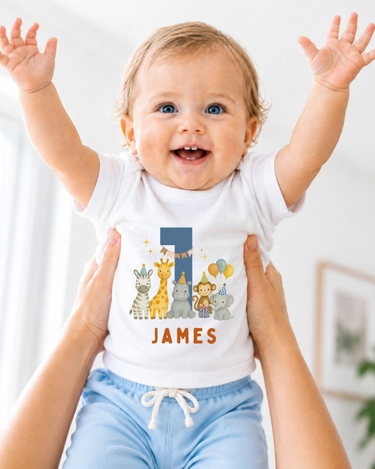 A smiling baby wearing a white onesie with a personalized number 1 and animal characters, being held up by someone's arms.