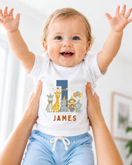 A smiling baby wearing a white onesie with a personalized number 1 and animal characters, being held up by someone's arms.