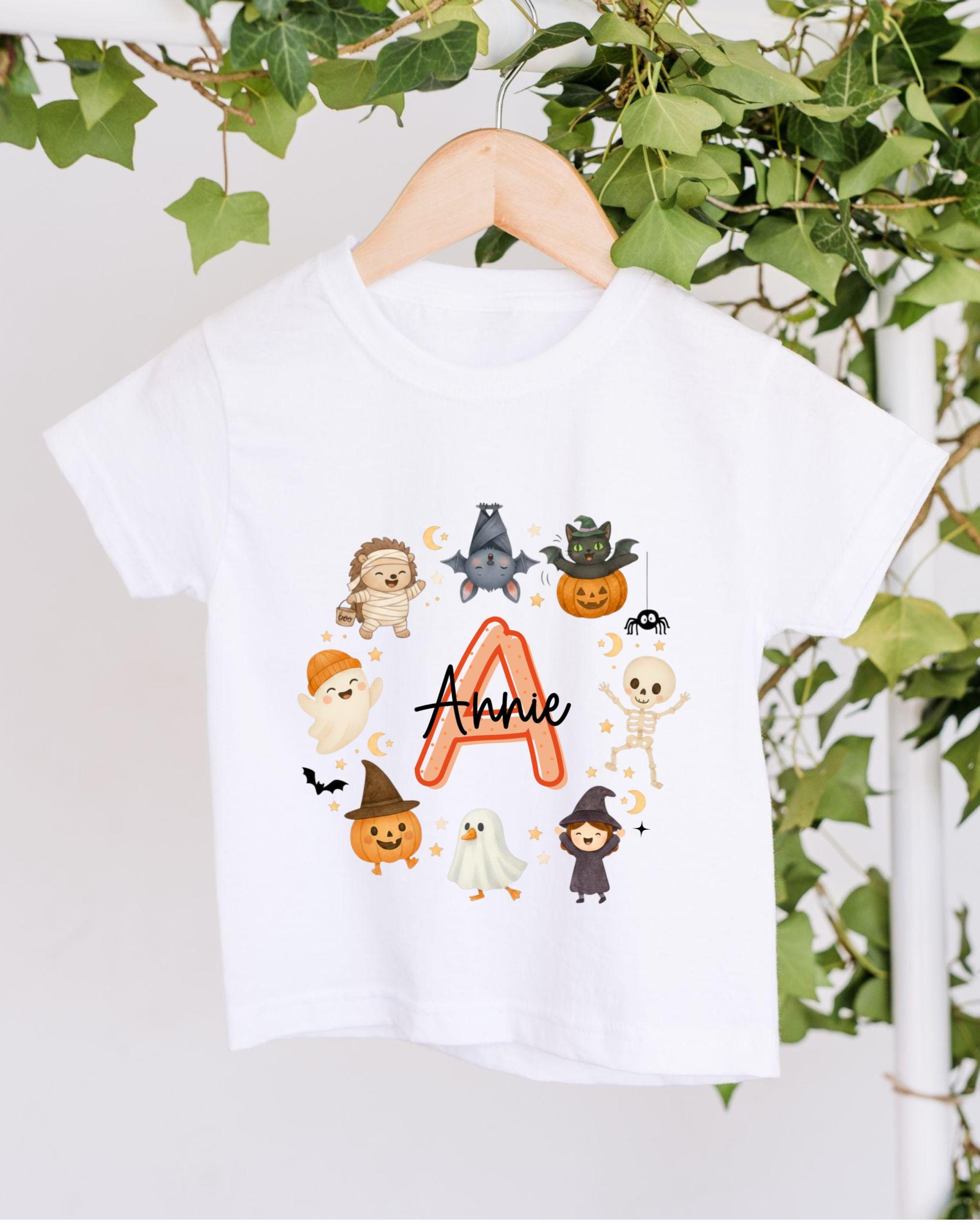 A white t-shirt with a graphic design featuring various cartoon-style characters, including a bat, skeleton, ghost, pumpkin, and a letter "A" surrounded by stars.