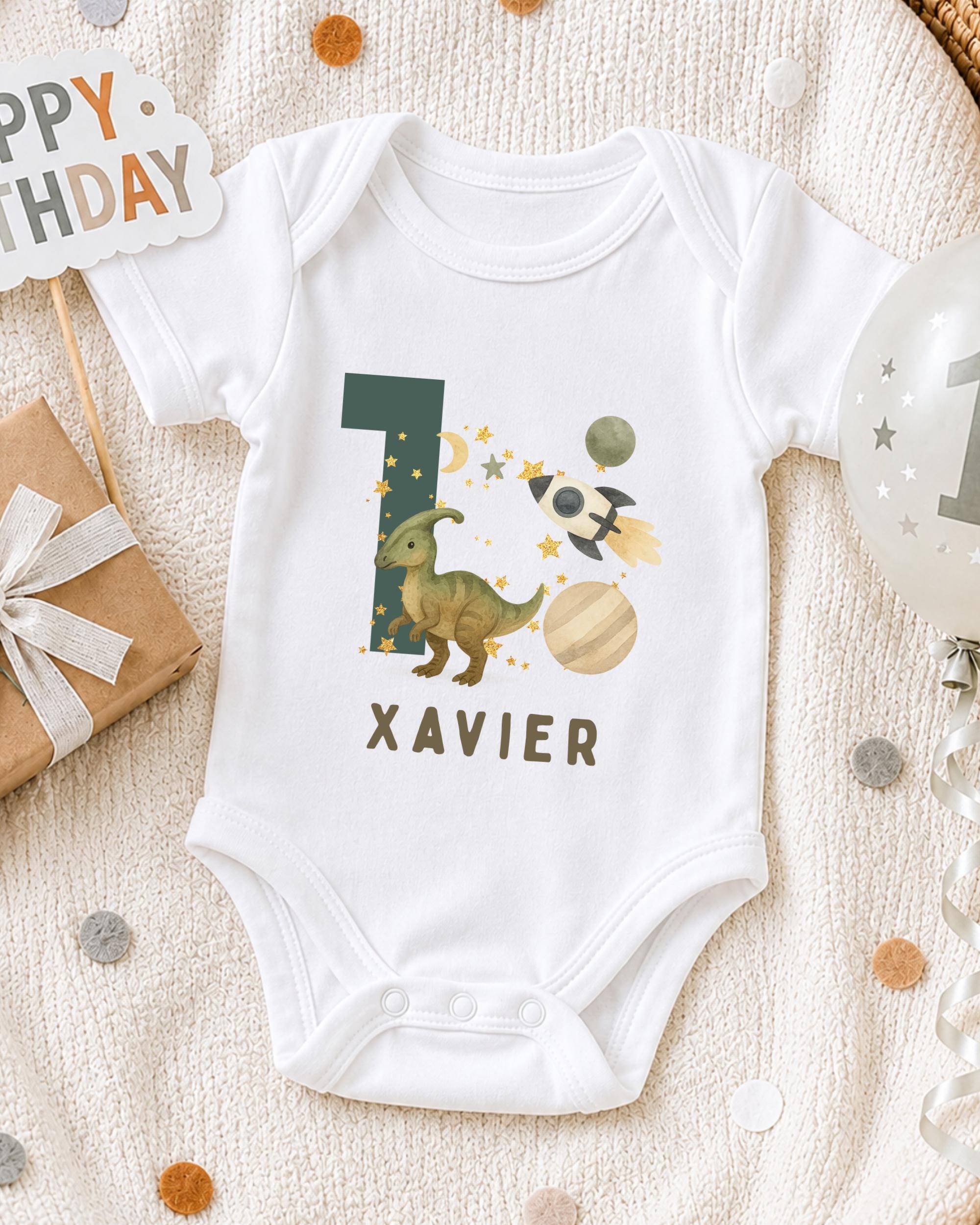 A white onesie with a dinosaur and space-themed design, along with a "XAVIER" name tag, is displayed on a bed with a "Happy Birthday" sign and a balloon.