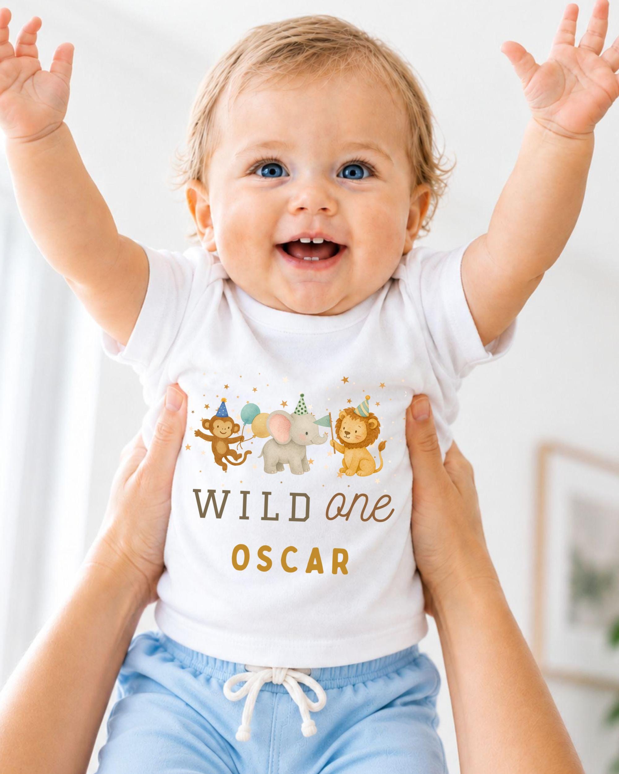 A smiling baby wearing a white t-shirt with a cartoon animal design and the text "Wild one Oscar" is being held up by two people.
