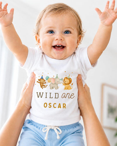 A smiling baby wearing a white t-shirt with a cartoon animal design and the text "Wild one Oscar" is being held up by two people.