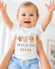 A smiling baby wearing a white t-shirt with a cartoon animal design and the text "Wild one Oscar" is being held up by two people.