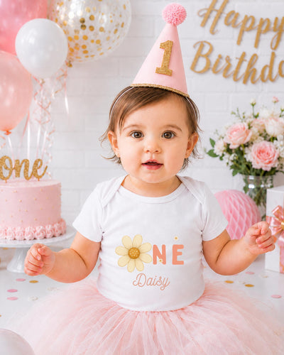 A young girl wearing a white t-shirt with a flower design and a pink birthday hat sits in front of a birthday cake and balloons, with a "Happy Birthday" sign in the background.