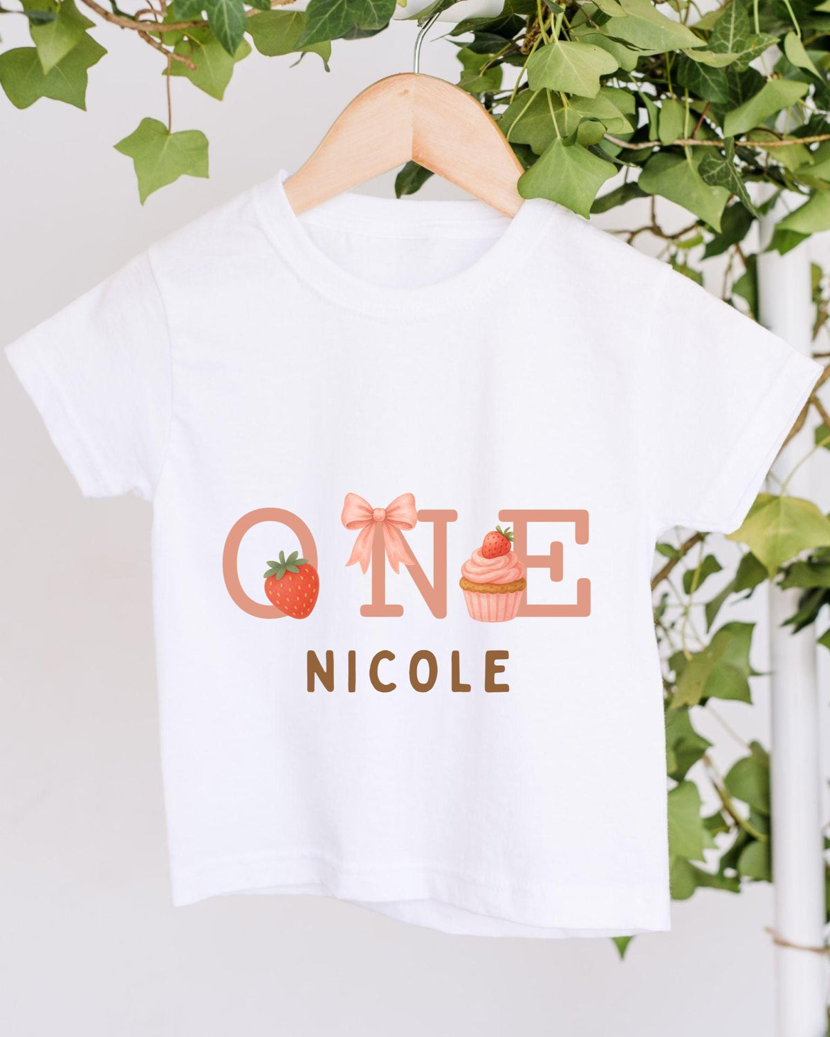 A white t-shirt with a graphic design featuring the text "ONE NICOLE" and illustrations of a strawberry and a cupcake.