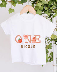 A white t-shirt with a graphic design featuring the text "ONE NICOLE" and illustrations of a strawberry and a cupcake.