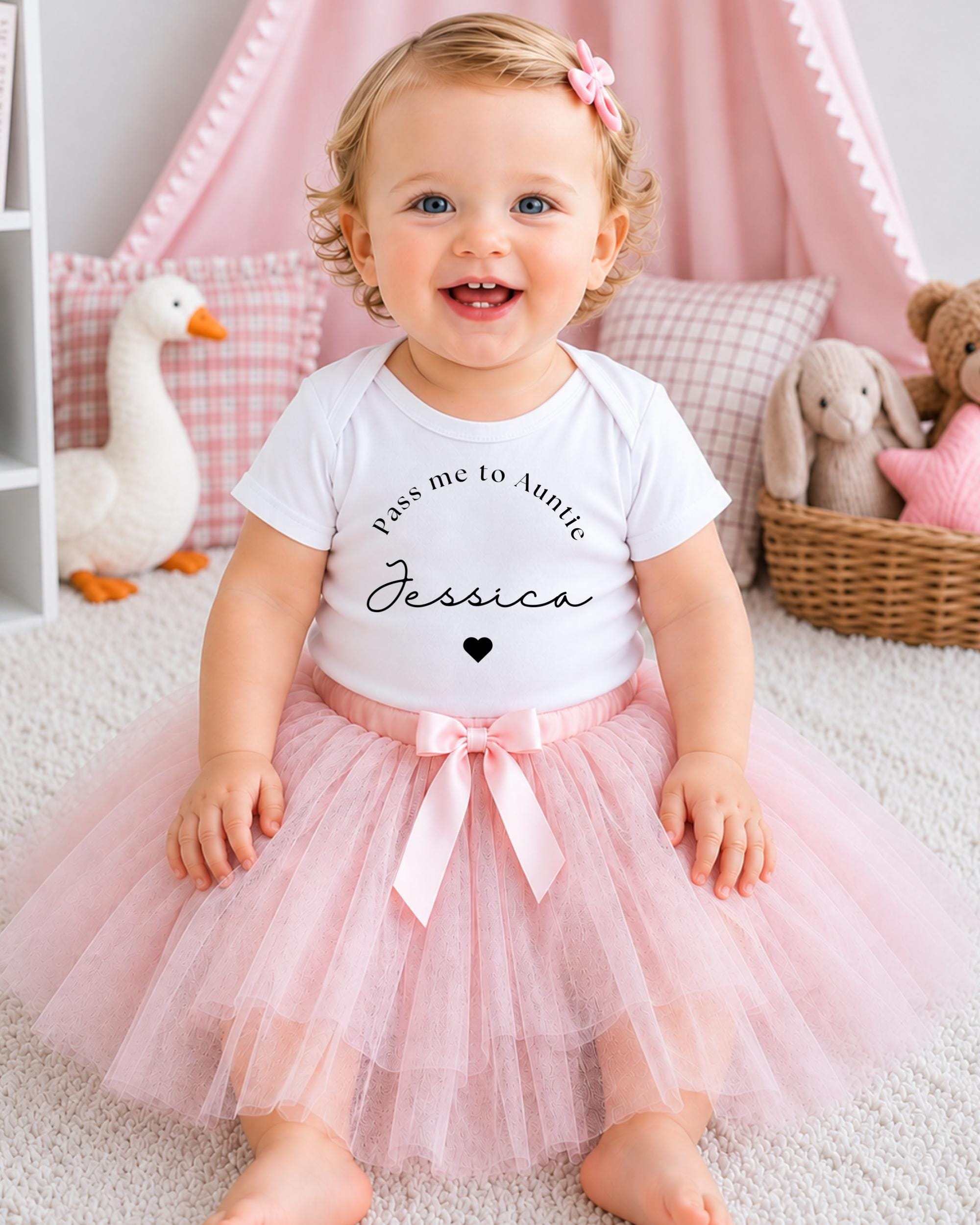 A smiling baby girl wearing a white t-shirt with the text "Pass me to Aunt Jessica" and a pink tutu skirt, sitting on a white carpet surrounded by stuffed animals.