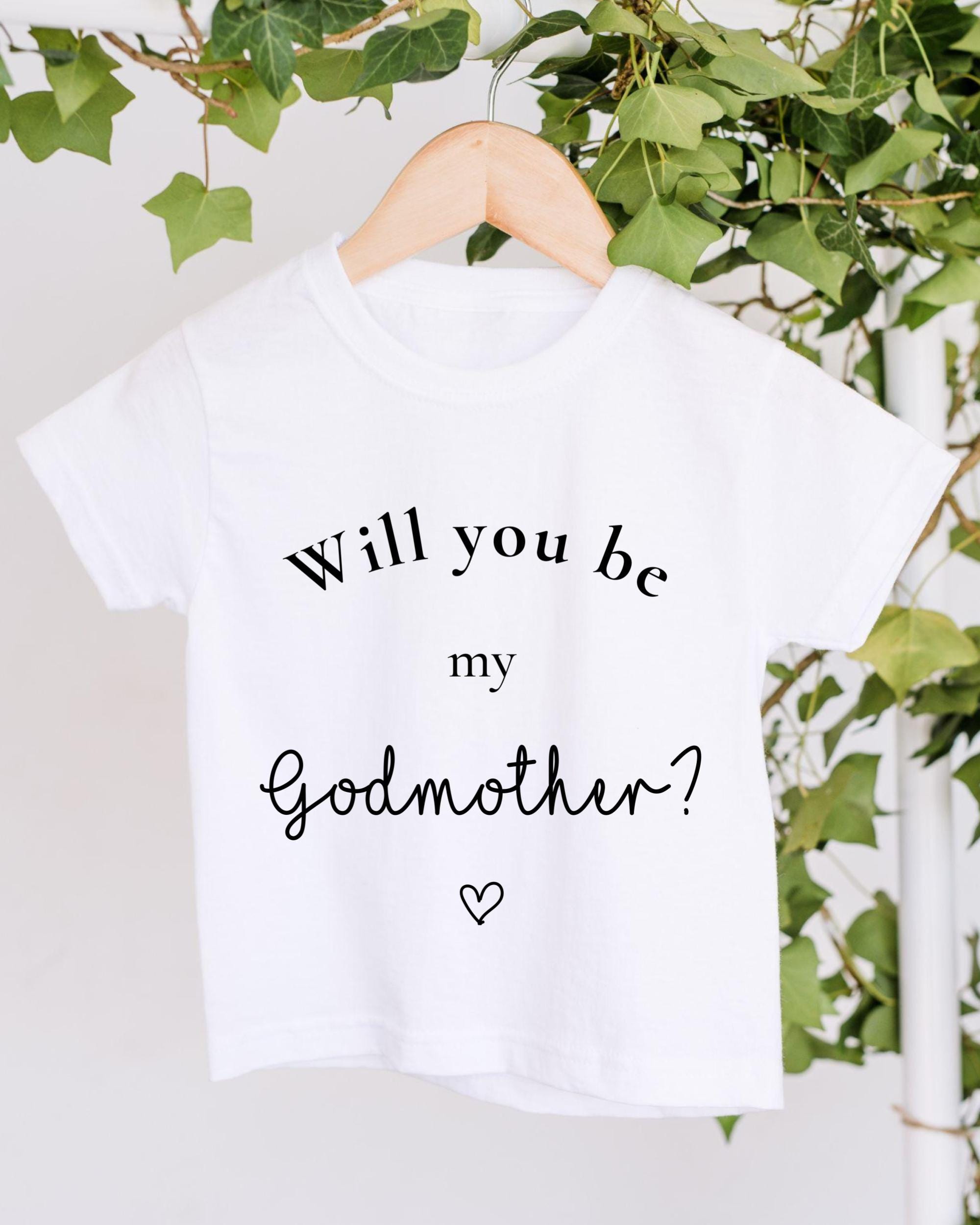 A white t-shirt with the text "Will you be my godmother?" printed on it, hanging on a wooden hanger against a backdrop of greenery.