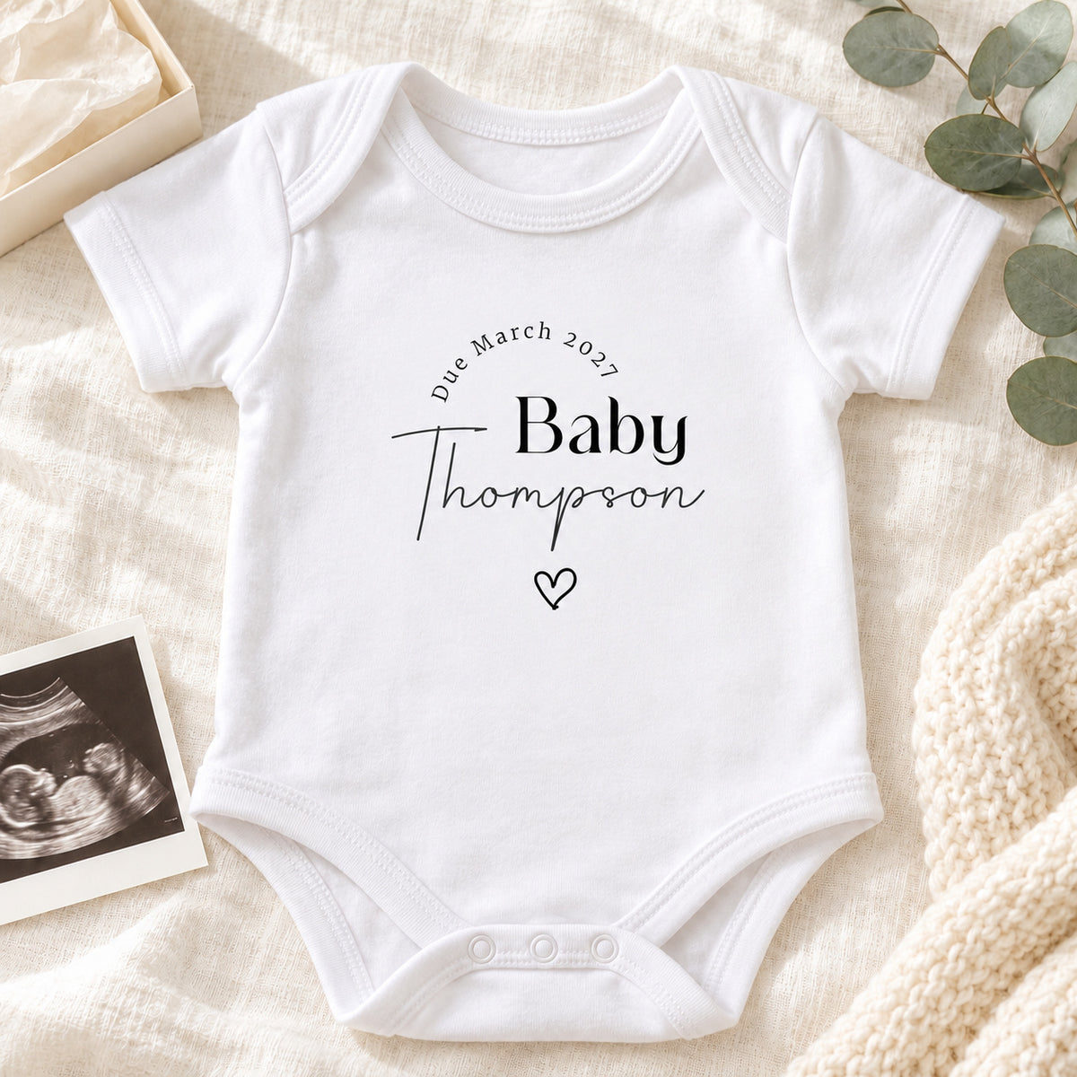 White personalised due date pregnancy announcement Baby Vest reading Due March 2027, Baby Thompson, styled with a cream gift box, ultrasound photograph and eucalyptus on beige linen, with props notice.