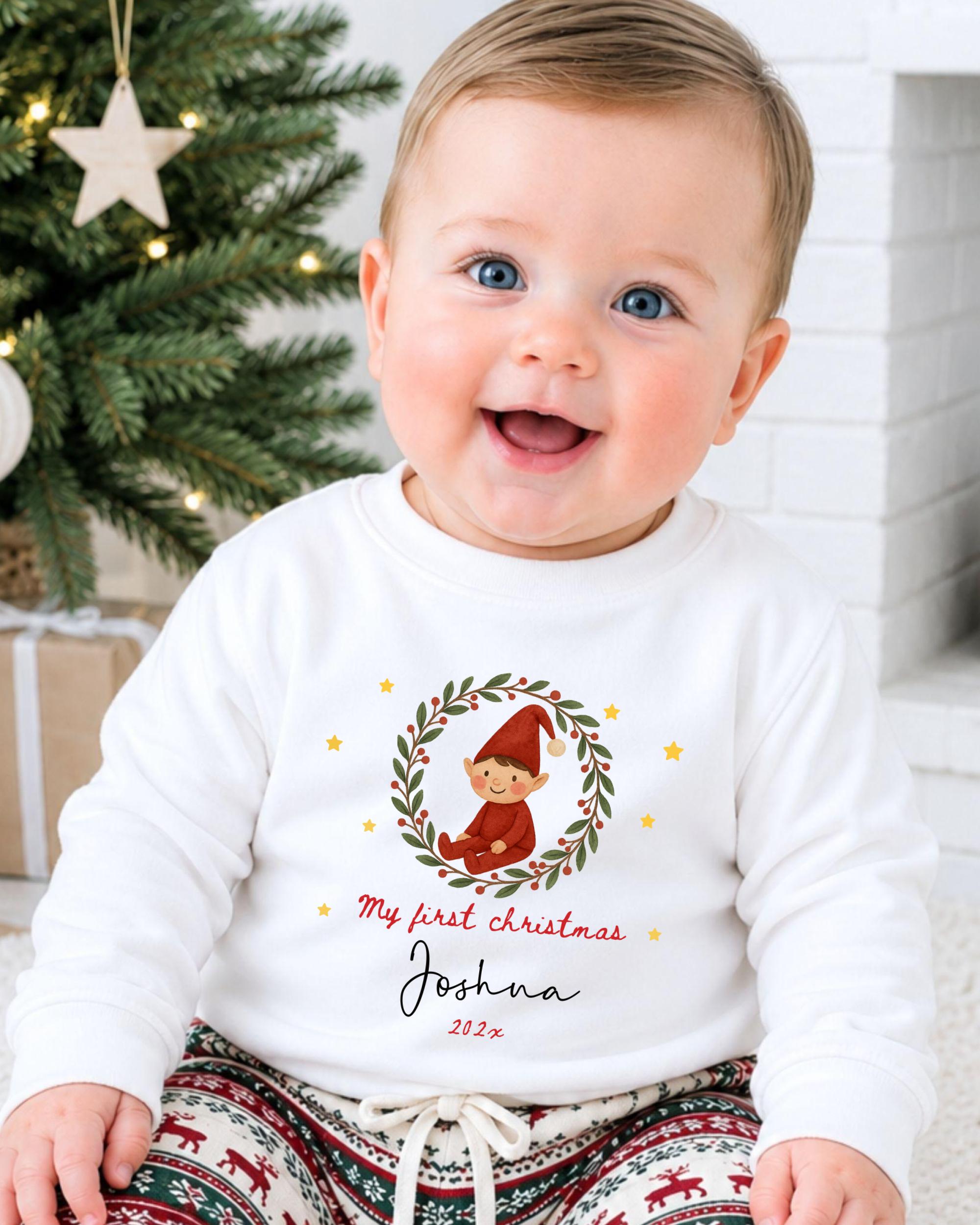A smiling baby boy wearing a white sweater with a Christmas-themed design and text, sitting in front of a Christmas tree.
