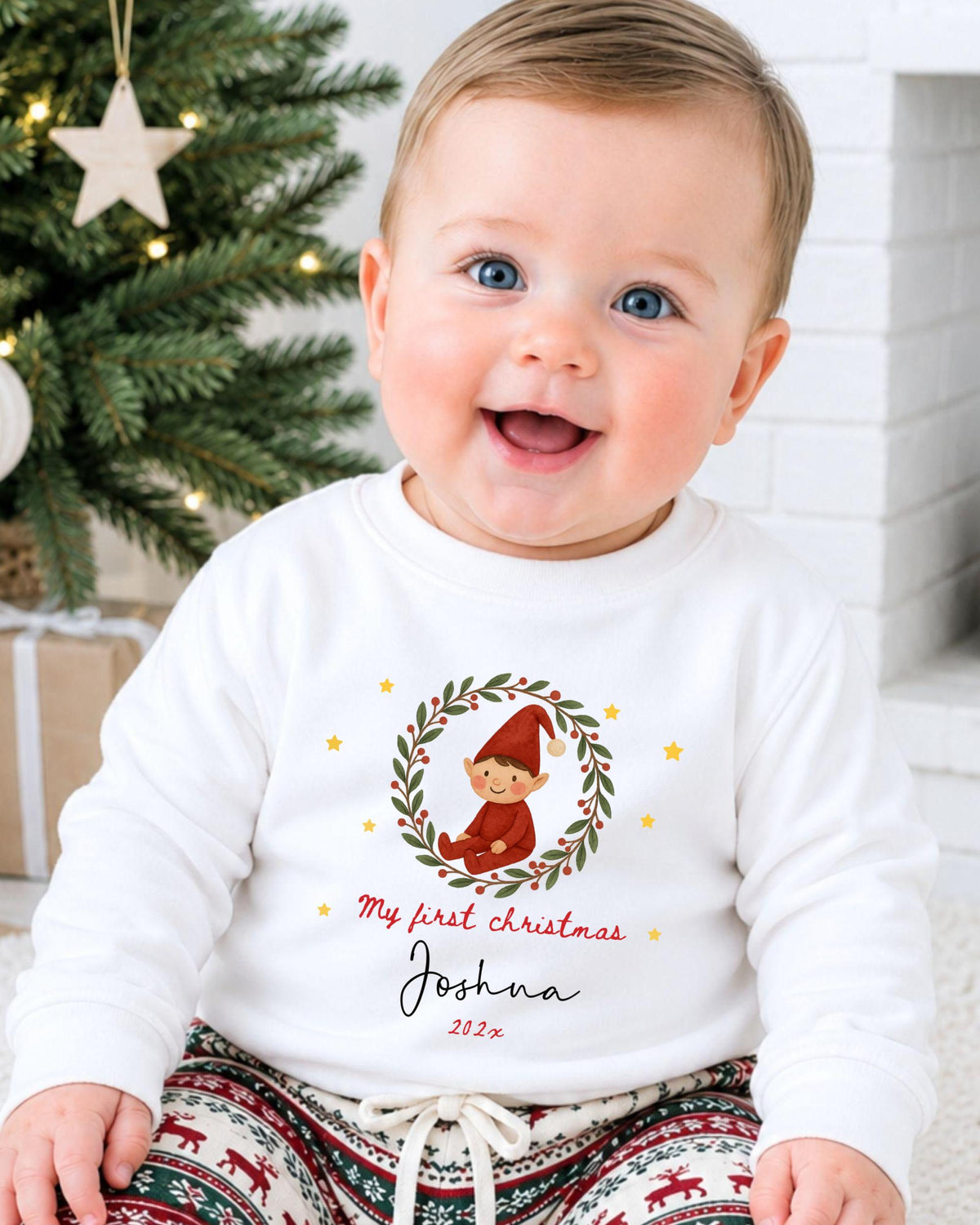A smiling baby boy wearing a white sweater with a Christmas-themed design and text, sitting in front of a Christmas tree.