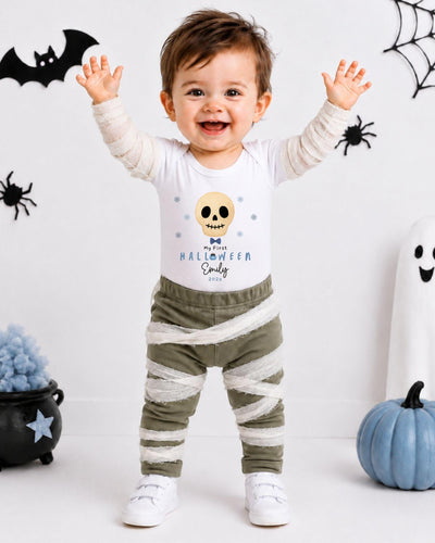 A young child wearing a Halloween-themed shirt and striped pants stands with their arms raised, smiling and surrounded by Halloween decorations.