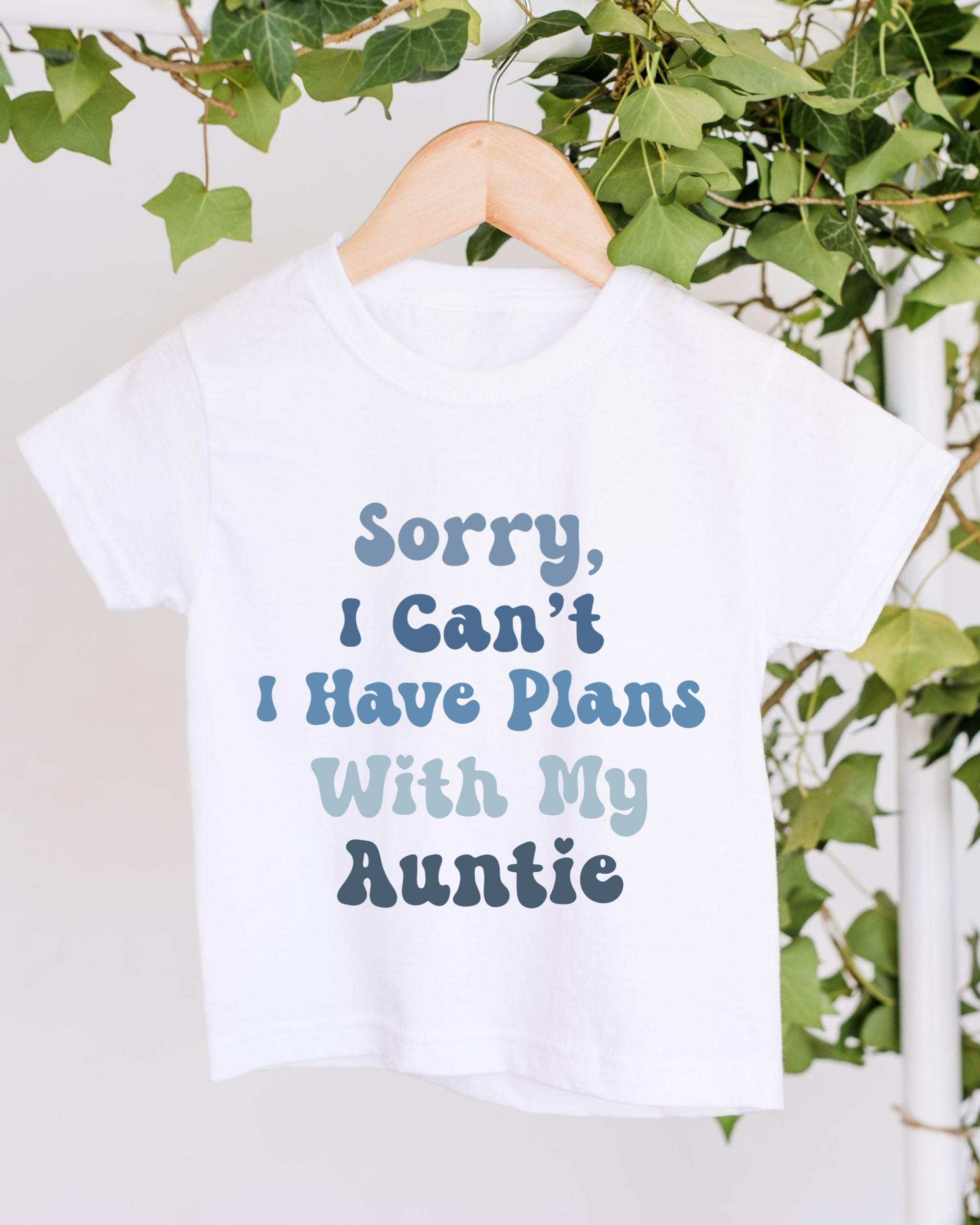 A white t-shirt with a humorous text printed on it, hanging on a wooden hanger.