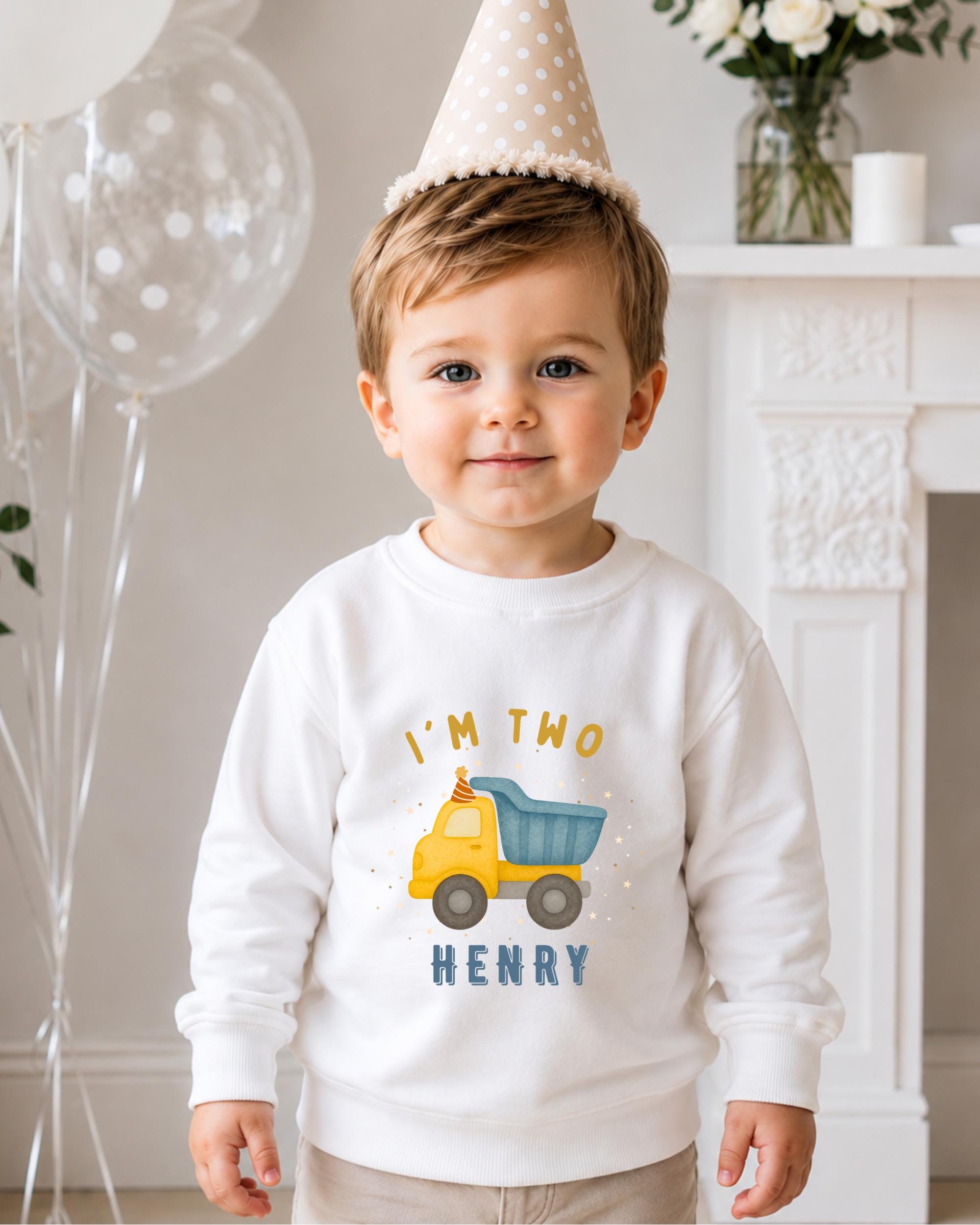 A young boy wearing a birthday hat and a white sweatshirt with a truck graphic and the text "I'm two Henry" is standing in front of a fireplace.