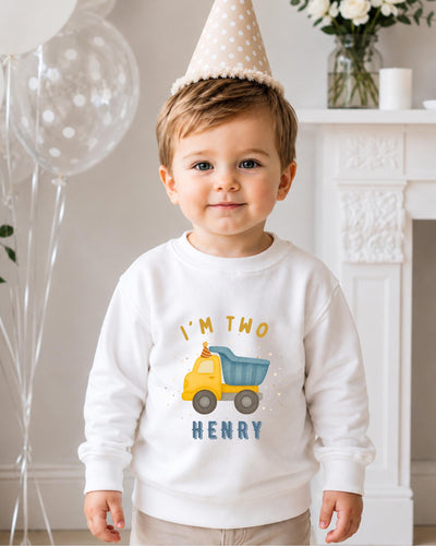 A young boy wearing a birthday hat and a white sweatshirt with a truck graphic and the text "I'm two Henry" is standing in front of a fireplace.