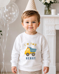 A young boy wearing a birthday hat and a white sweatshirt with a truck graphic and the text "I'm two Henry" is standing in front of a fireplace.