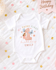 A white onesie with a fairy and butterflies printed on it, placed on a bed with confetti and a birthday card.
