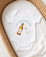 A white onesie with a graphic design on it, featuring a bottle of beer and a baby bottle, along with the text "DADMY'S NEW DRINKING BUDDY".