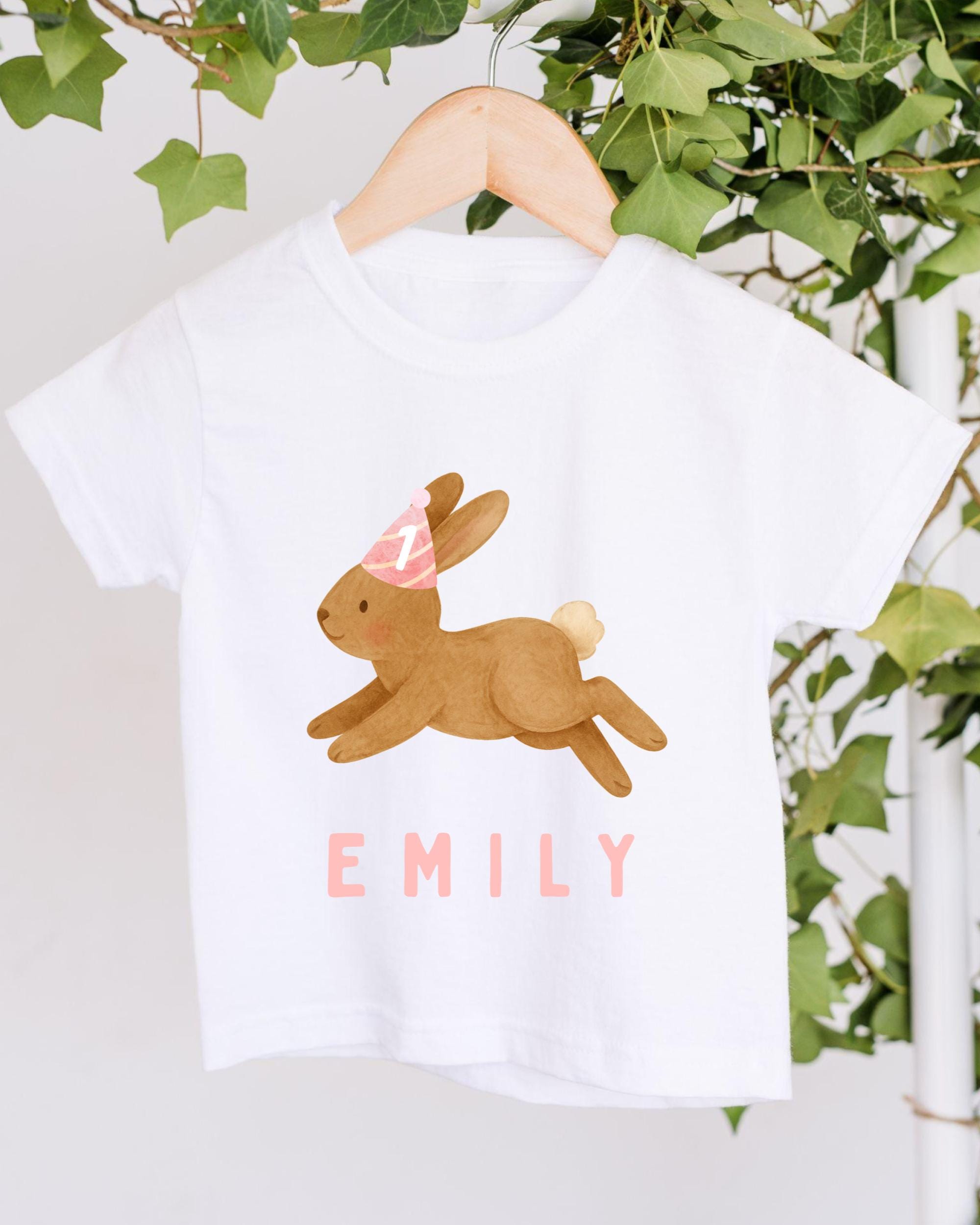 A white t-shirt with a cartoon image of a bunny wearing a party hat and the name "EMILY" printed on it, hanging on a wooden hanger.