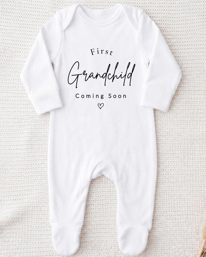 A white bodysuit with the text "First Grandchild Coming Soon" printed on it.