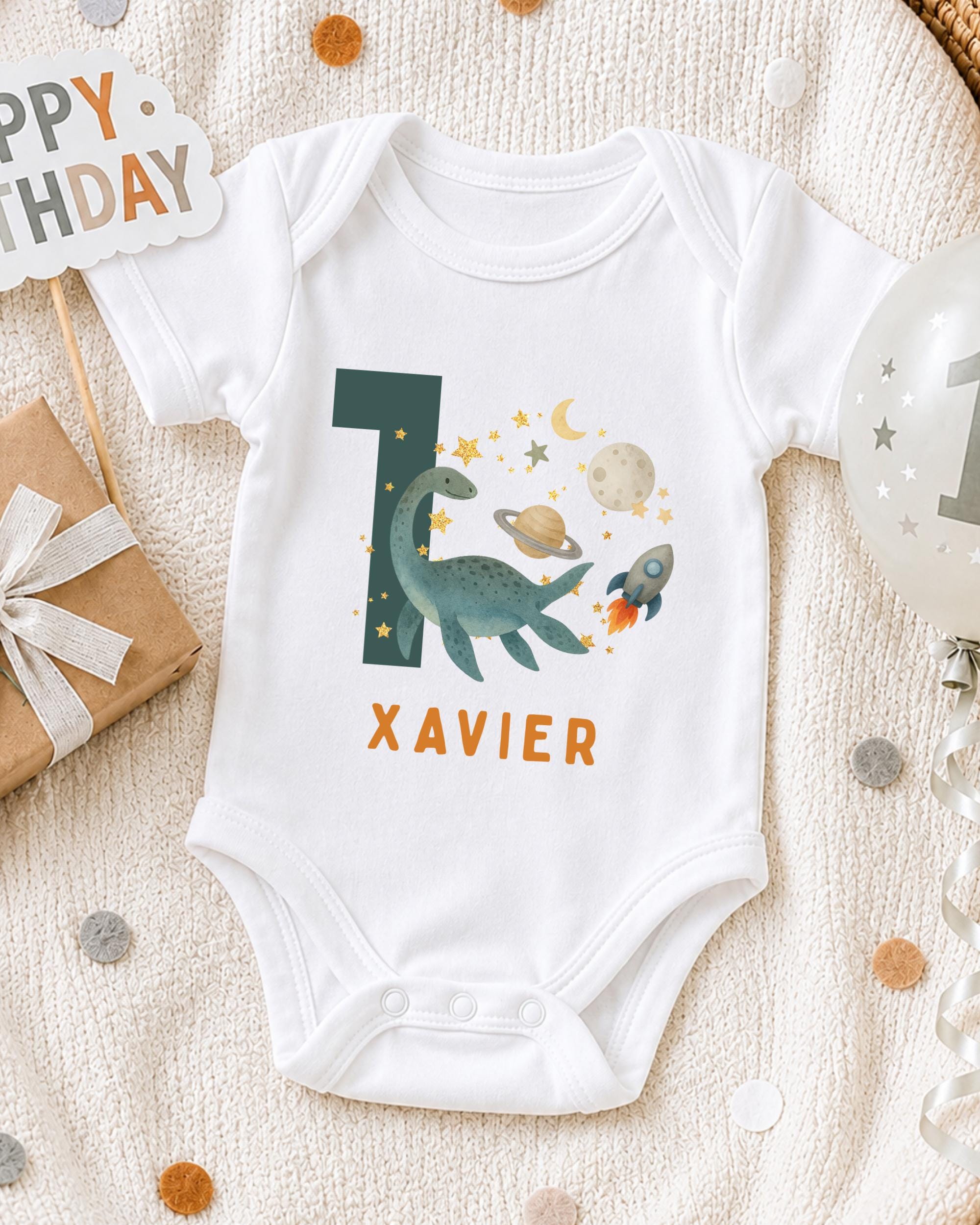 A white onesie with a dinosaur and space-themed design, along with a "XAVIER" name tag, is displayed on a bed with a "Happy Birthday" sign and a balloon.