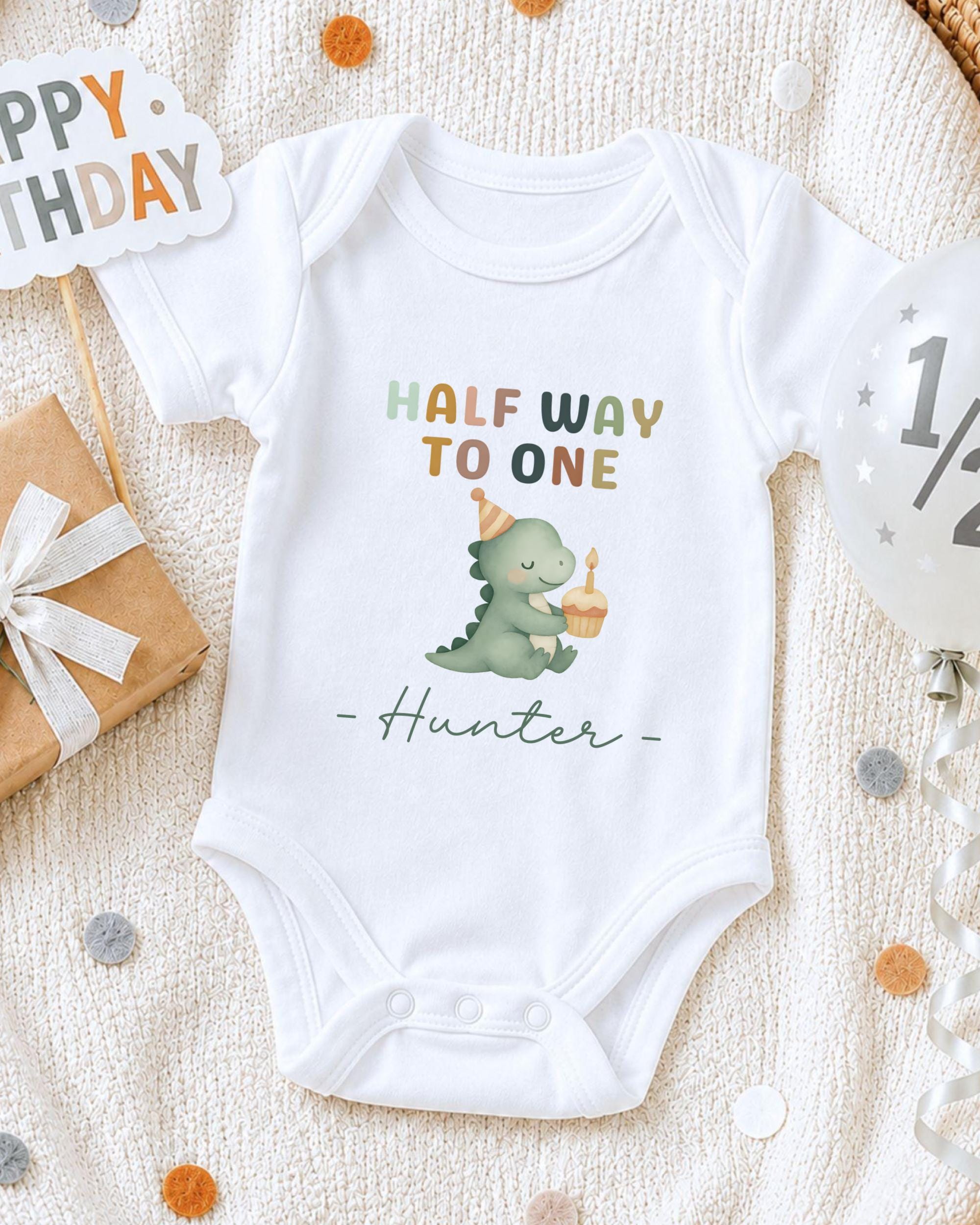 A white onesie with a cartoon dinosaur and the text "Half Way to One Hunter" printed on it, along with a birthday sign and confetti.