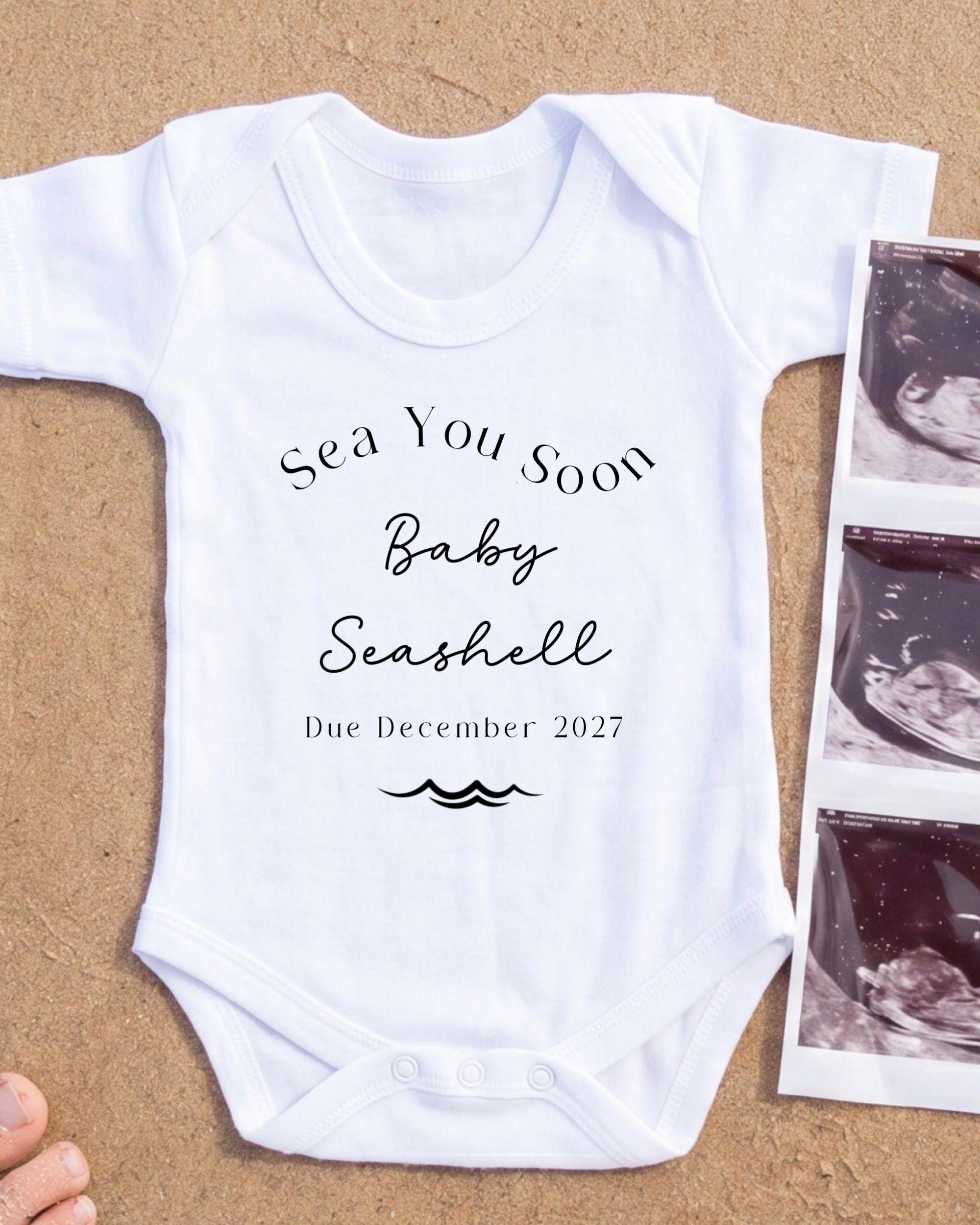 A white onesie with text and a graphic related to the announcement of a baby's arrival.