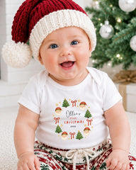A smiling baby wearing a Santa hat and a white t-shirt with a Christmas-themed design.