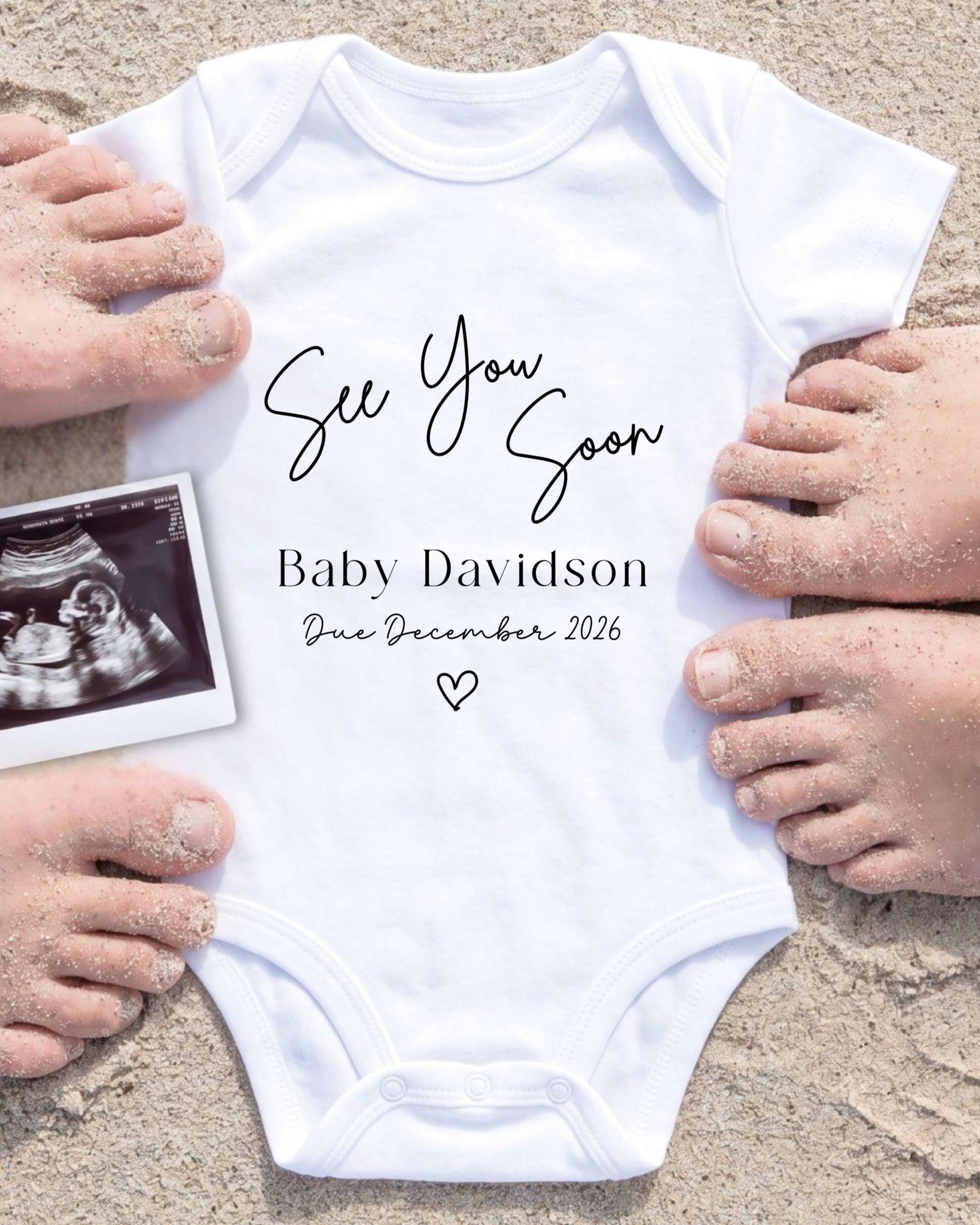 a white baby onesie with the text "See You Soon Baby Davidson Due December 2026" printed on it, surrounded by sand and a black and white photograph of a baby's belly.
