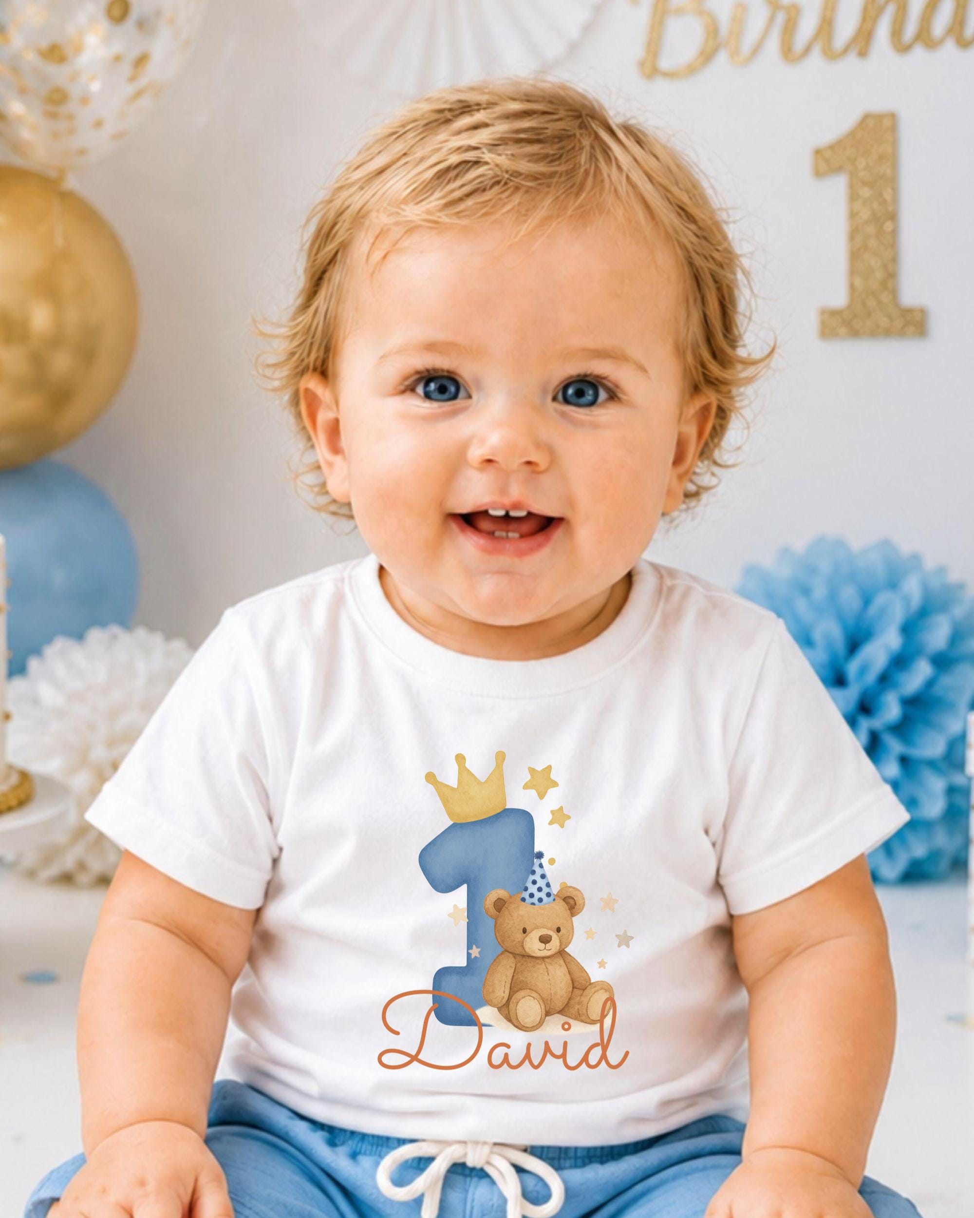 A smiling baby boy wearing a white t-shirt with a teddy bear and a number one, sitting in front of a birthday party backdrop.