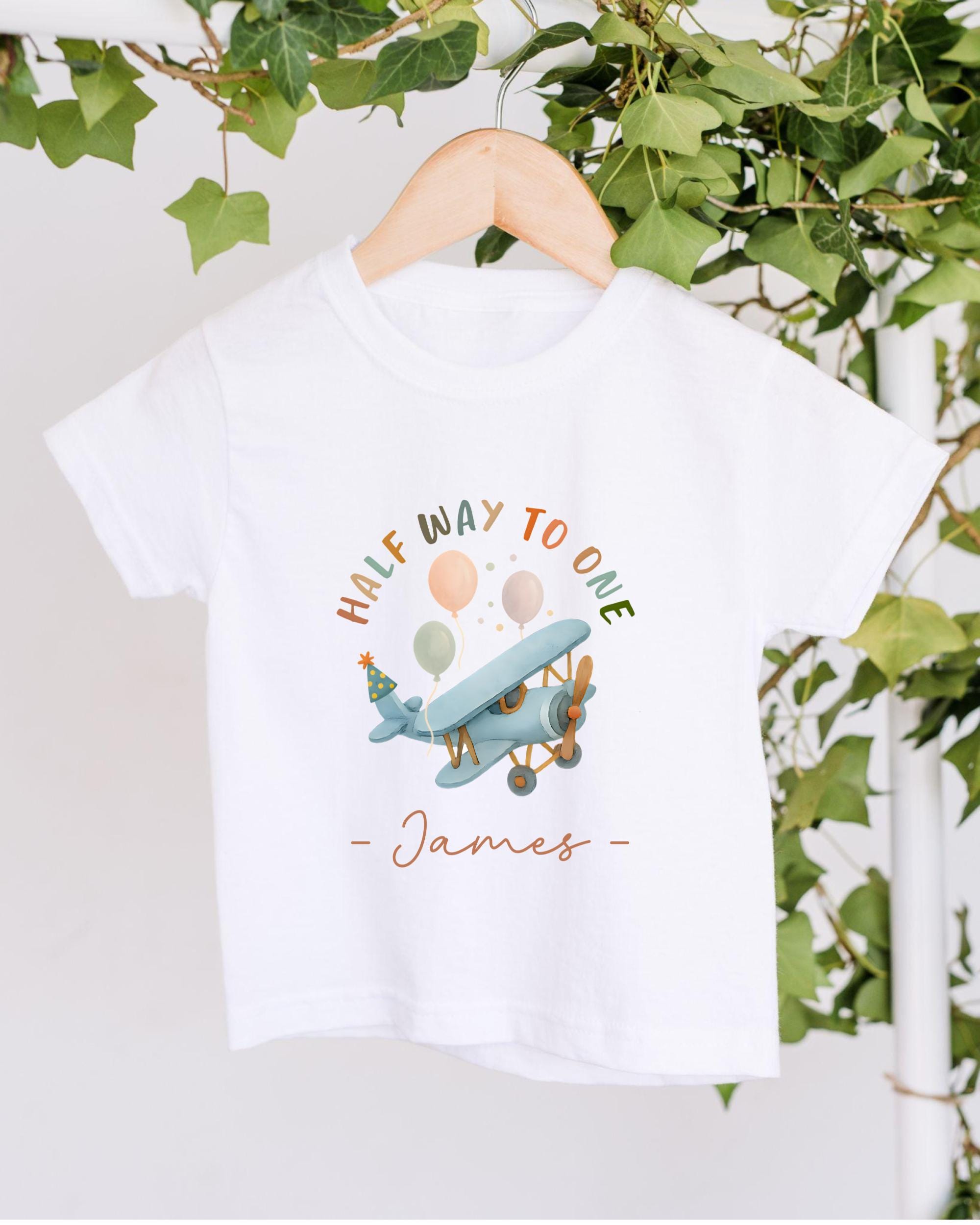 A white t-shirt with a graphic design of a blue airplane surrounded by colorful balloons, hanging on a wooden hanger.