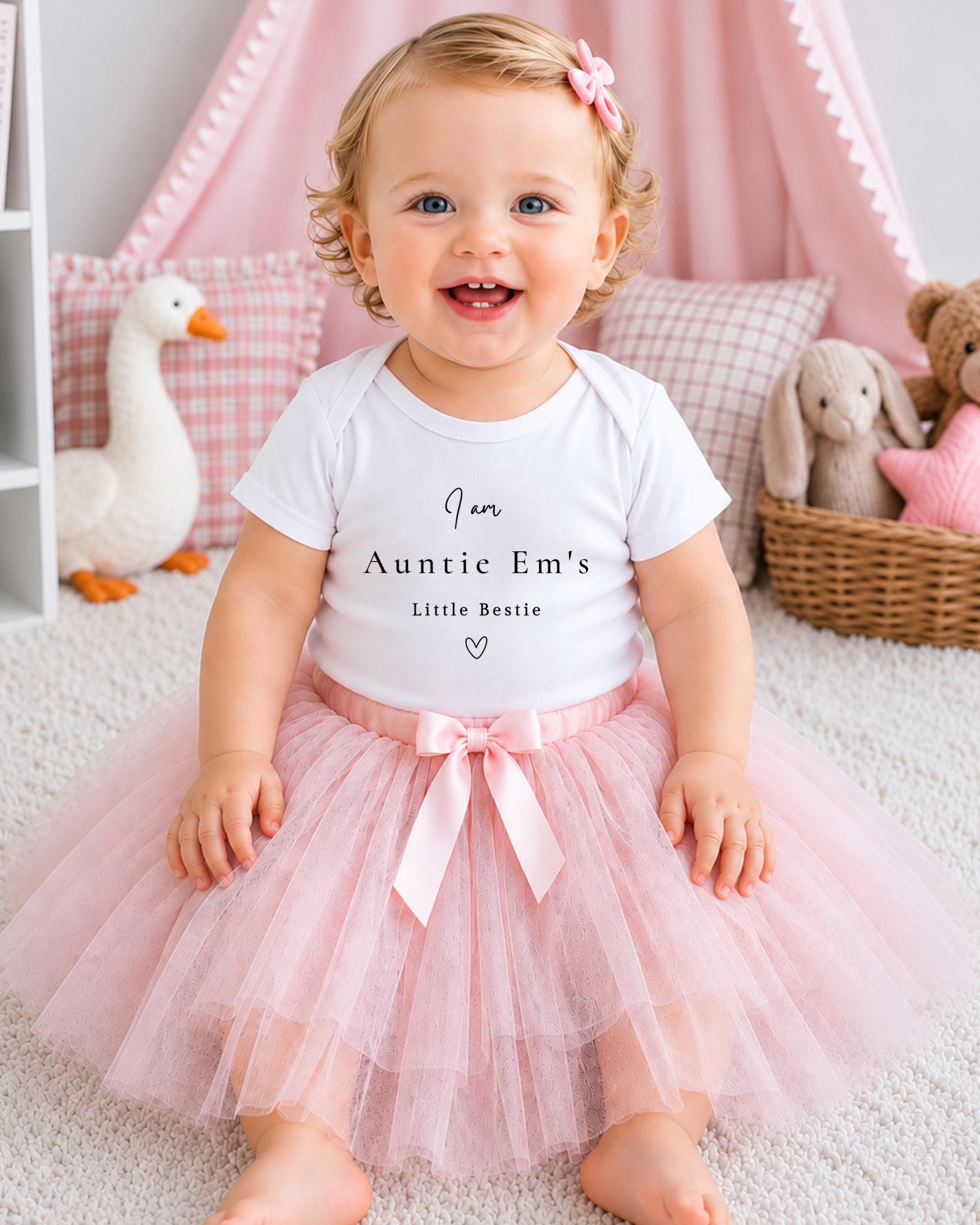 A smiling baby girl wearing a white t-shirt with text and a pink tutu sits on a white carpet, surrounded by stuffed animals and a pink canopy.