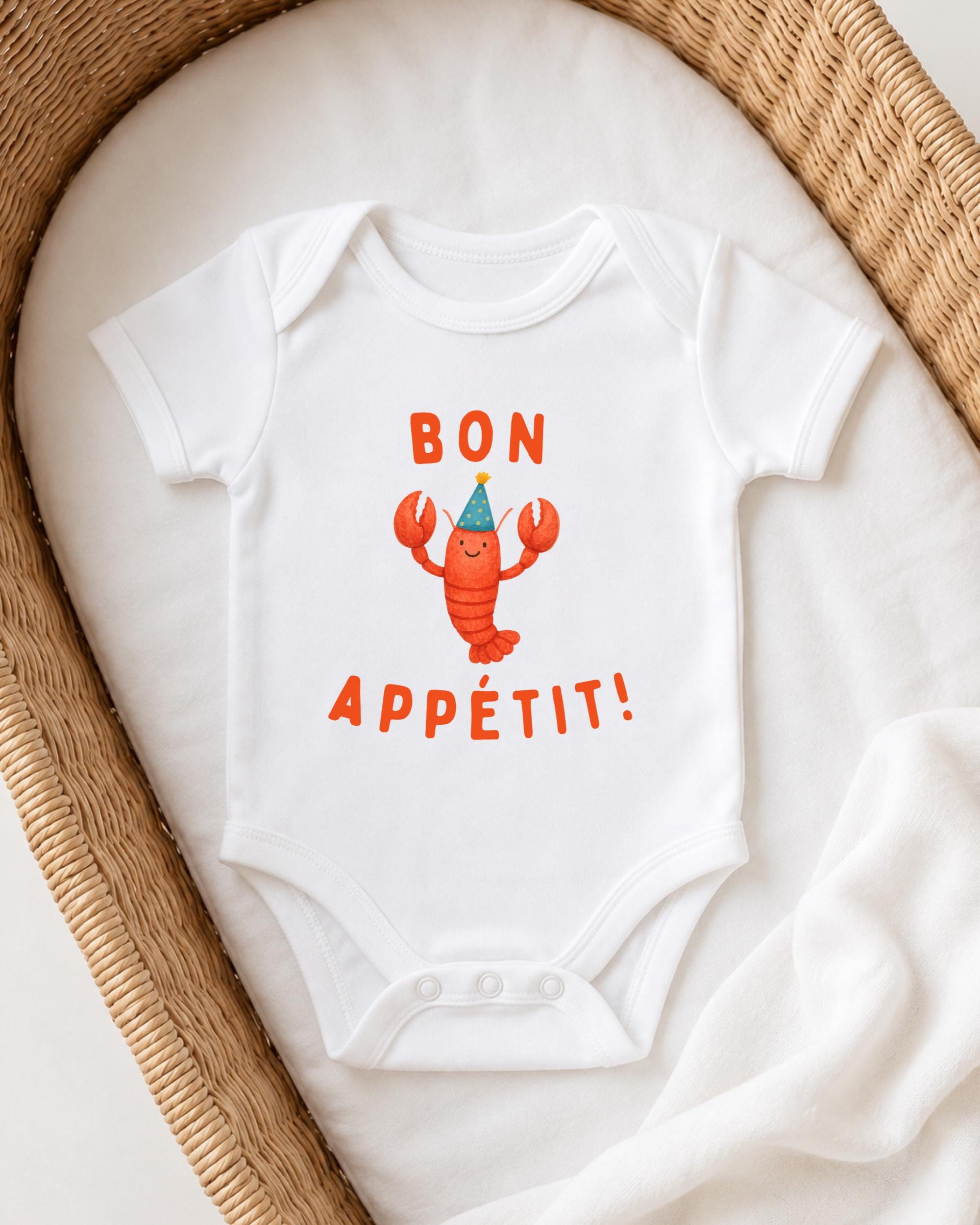 A white onesie with a cartoon lobster and the text "BON APETIT!" printed on it, placed on a white blanket.