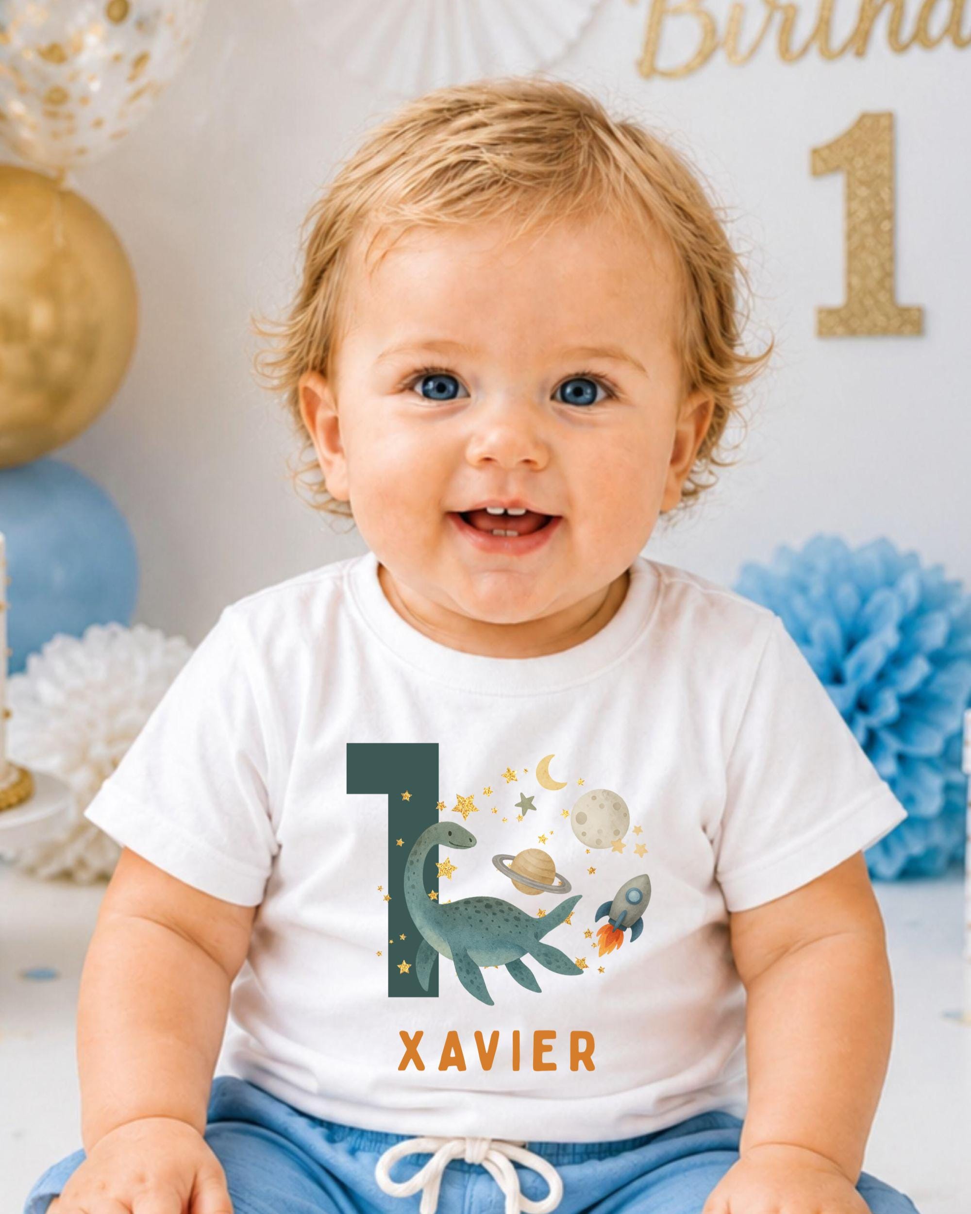 A smiling baby boy wearing a white t-shirt with a dinosaur and space-themed design, sitting in front of a birthday party backdrop.
