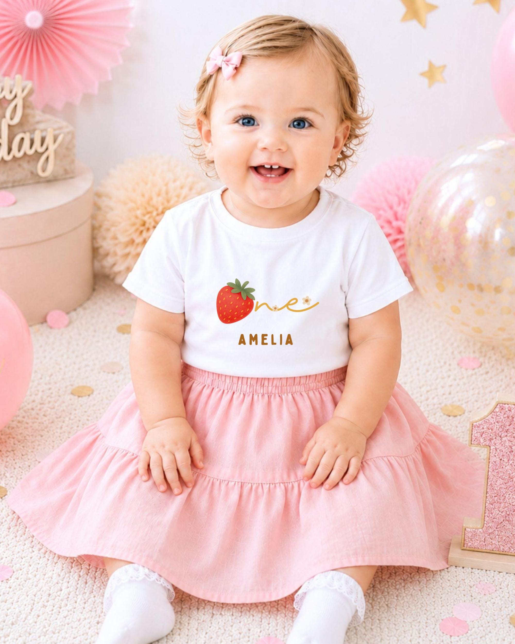 A smiling baby girl wearing a white t-shirt with a strawberry design and a pink skirt, sitting on a pink and white background with balloons and confetti.