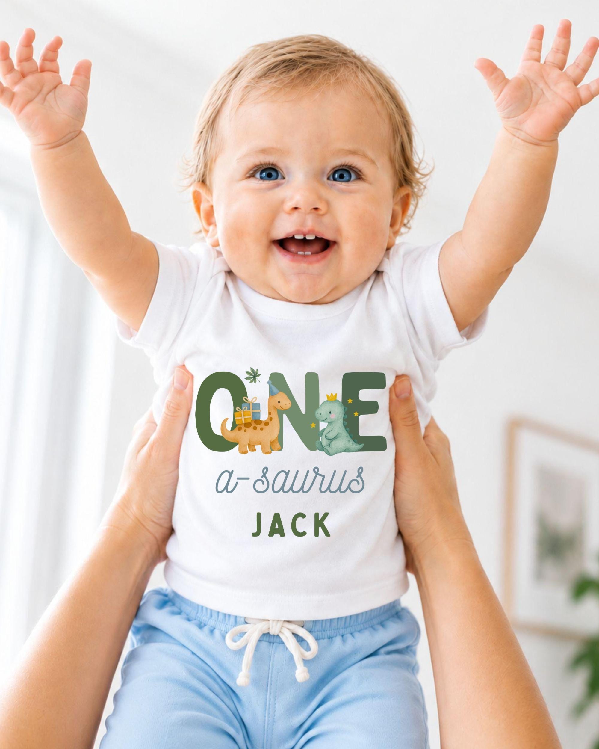 A smiling baby wearing a white shirt with a dinosaur graphic and the text "ONE a-saurus JACK" is being held up by someone's arms.