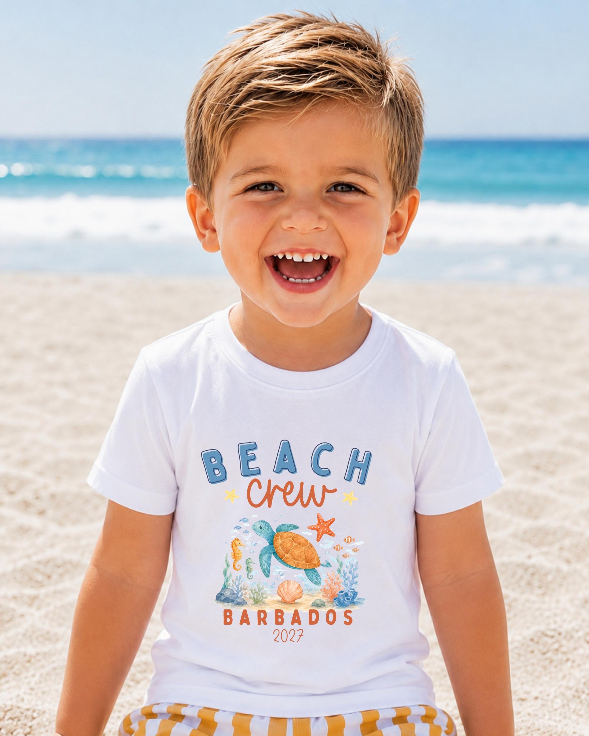 A young boy with a big smile on his face, wearing a white t-shirt with a beach crew design and text, standing on a sandy beach with the ocean in the background.