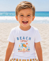 A young boy with a big smile on his face, wearing a white t-shirt with a beach crew design and text, standing on a sandy beach with the ocean in the background.